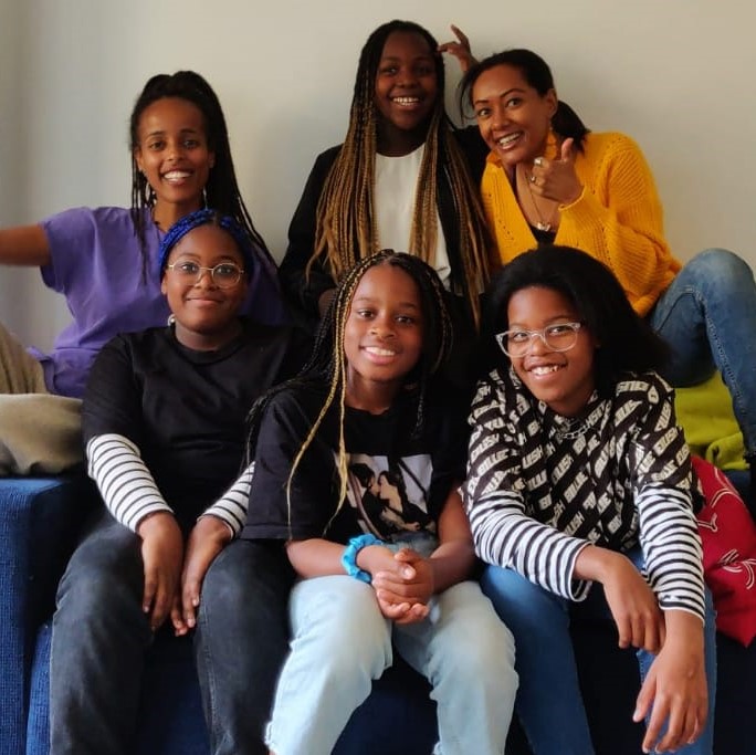 Kuusi afrikkalaistaustaista adoptoitua tyttöä ja nuorta naista hymyilee. 